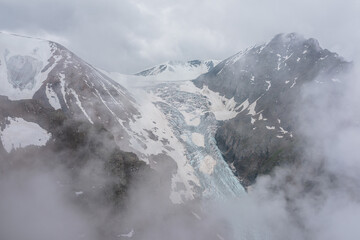 Atmospheric mountain landscape with long vertical glacier tongue with cracks among rocks in dense low clouds. Awesome aerial view to large glacier with icefall in thick low clouds in high mountains.
