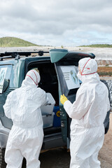 Rear viiew of environmental scientists in hazmat suits and masks standing at car and finding tool in container