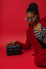 Fashionable Black woman posing with trendy quilted leather bag. Model wearing classic red suit with...