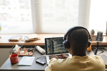 Rear view of black man in wireless headphones typing on laptop while programming code on software
