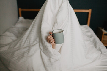 Pretty young woman sits in bed wrapped in a blanket with a cup of coffee