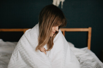 Pretty young woman sits in bed wrapped in a blanket