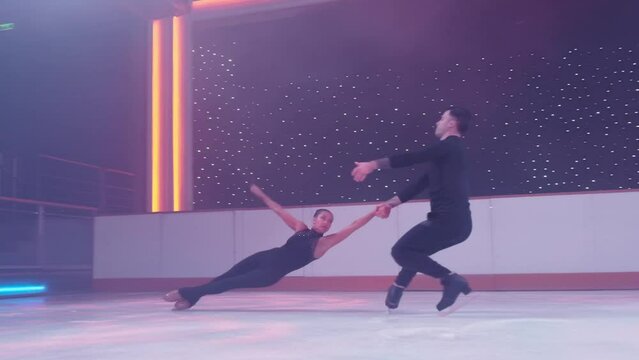 Pair of figure skaters rotating on ice rink,woman goes into death spiral,hovers in air parallel to ice her legs in split, man holds her by hands. Figure skating lift,slow motion support,distant shot.