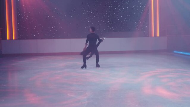 Figure skaters pair on ice performs stationary rotational support,prohibited element. Woman in split upside down,hips angle 180, man in crouch holding her with straight hands by arm,leg.Slow motion.