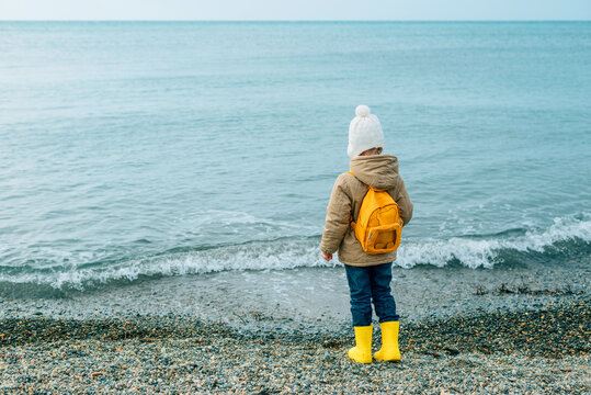 Little Girl Runs Along The Rocky Coast Of The Sea In Autumn In Outerwear And Rubber Boots, Back. A Child Runs Along The Beach Of The Sea, Slapping The Water With His Feet