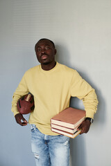 Portrait of serious African-American student guy standing with rugby ball and stack of books against gray wall