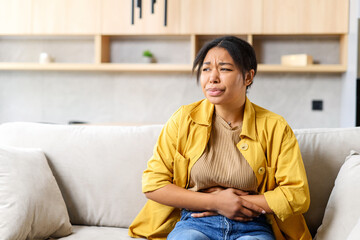 Young African-American woman sitting on the couch and suffers from month menstruation period pain,...