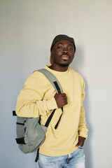 Portrait of serious handsome Black student guy in yellow sweatshirt and cap standing with satchel against isolated baclground