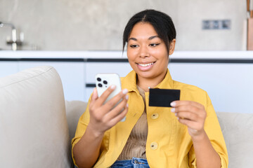 Cheerful young woman using smartphone shopping app for buying online, holding credit card and...