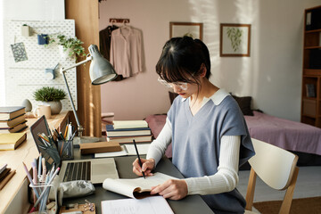 Serious Asian girl in eyeglasses sitting at table and preparing for seminar in campus room