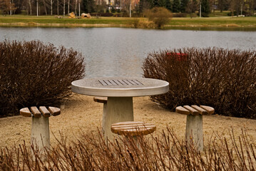 Parkowy stolik do szachów ( i nie tylko ) wraz z siedziskami , wśród krzewów ozdobnych , nad stawem wczesną wiosną .  - obrazy, fototapety, plakaty