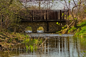 Kamienny mostek z drewnianymi poręczami wśród drzew  , wczesną wiosną nad małą rzeczką ( strumieniem ) .