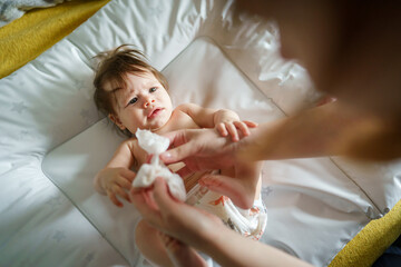 One small caucasian baby lying on the bed naked with hands of unknown woman mother using wet wipe...
