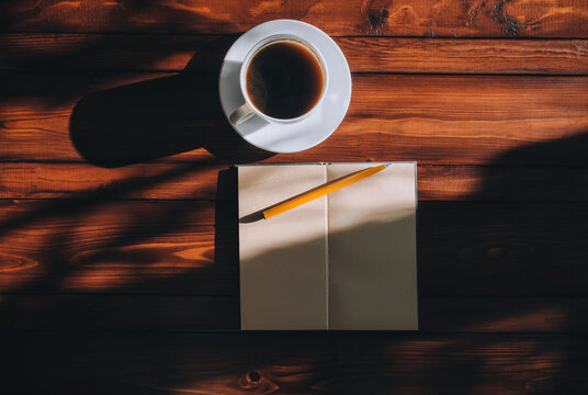 A paper notebook, a pen and a cup of coffee lie on a brown mahogany wooden background with patches of sunlight. Copy space, layout. The concept of loneliness, journalism and writing.
