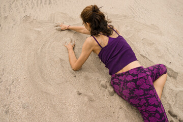 Latin woman wearing a purple leggings and top sitting on the sand at the beach and practicing a yoga movement  by twisting her body. Summertime and Relax concept. Copy space.