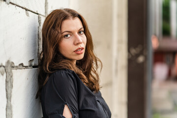 Fototapeta premium Front view portrait of adult caucasian woman posing in front of white wall outdoor in day looking to the camera wearing black dress and brown hair fashion