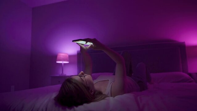 Girl laying on back browses social media on smart phone late at night in bedroom.