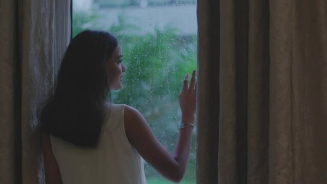 Close up shot of woman removing curtain to see rain from window in Diu city of India. Close up of a young woman is touching a glass with water drops and looking outside while is raining.