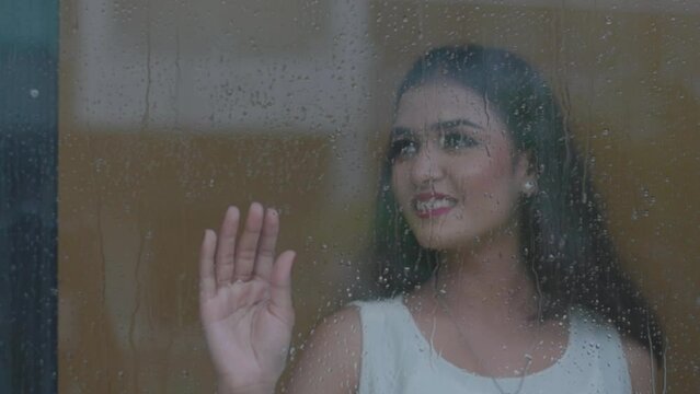 Close up portrait of girl standing near the window while its raining outside at Diu city of India.  Bored girl behind window in a rainy day. Shot in 4k