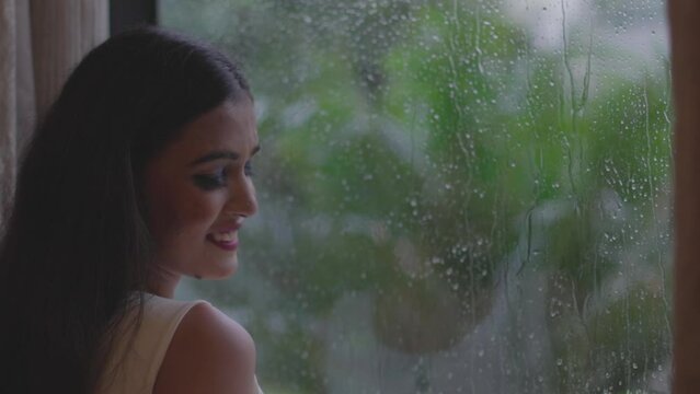 Young happy girl enjoying the rainy season seeing from window at Diu City of India. Happy joyful smiling girl woman is standing near the window. It is raining outside and the lights of the city.