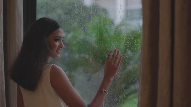 Close up shot of woman removing curtain to see rain from window in Diu city of India. Close up of a young woman is touching a glass with water drops and looking outside raining.