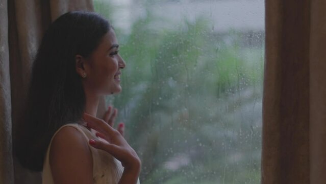 Back view of smiling woman seeing  outside rainy weather from window in Diu city of India. Close up of a young woman is touching a glass with water drops