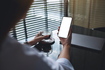 Mockup image of a woman holding mobile phone with blank desktop screen while drinking coffee with laptop on the table