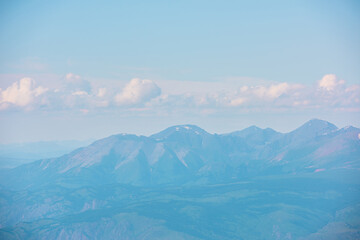 Minimalist aerial landscape with forest hills and high mountain range in sunlight and haze at changeable weather. Minimal alpine scenery with sunlit mountain tops silhouettes in mist under cloudy sky.