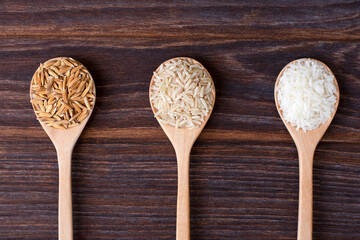 Various types of rice ; paddy rice, brown coarse rice and white thai jasmine rice in wooden spoon isolated on rustic wood board background. Flat lay. Top view.