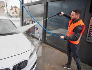 Young man washing white car on carwash station, wearing orange vest. Handsome worker cleaning automobile, using high pressure water.