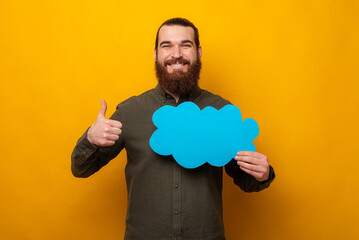 Cheerful bearded man is holding a blue cloud bubble speech and showing thumb up.