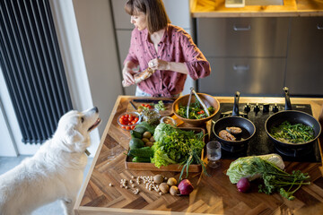 Young housewife makes a salad while standing with her adorable white dog in the kitchen. Table with...