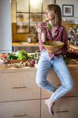 Portrait of a young cheerful woman dressed casually sitting on the table and eating healthy salad...