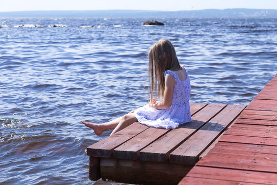 Little blonde girl in a white dress sitting on a wooden pier, surrounded by water, and enjoying the beautiful view of the lake in summer