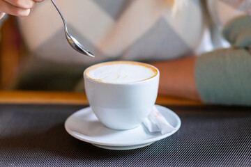 White cup of cappuccino on the background of a blurred teaspoon and female hands