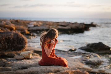 A European teenage girl in a dress sits on stones on the seashore, peace and quiet, a lonely child looks at the sea