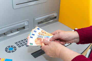 RETIRED SENIOR WOMAN WITHDRAWING EURO BANKNOTES FROM AN ATM. DIGITAL DIVIDE FOR THE ELDERLY.