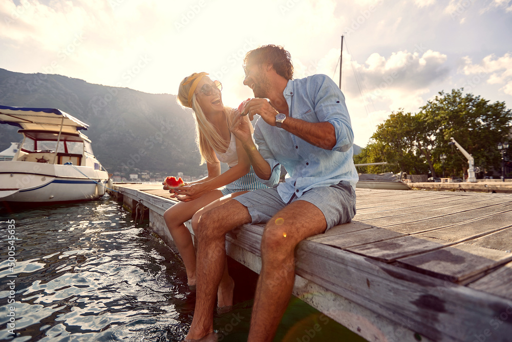 Wall mural lovely young couple sharing moments of bliss and eating watermelon sitting on wooden jetty by water.