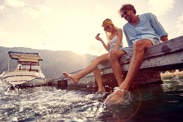Playful couple sitting on wooden jetty by water, eating watermelon and making splashes in water. ...