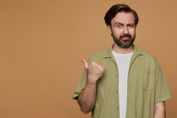 studio portrait of bearded man posing over beige background with shocked facial expression, points...