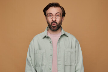 studio portrait of bearded man posing over beige background looking into camera with shocked facial expression