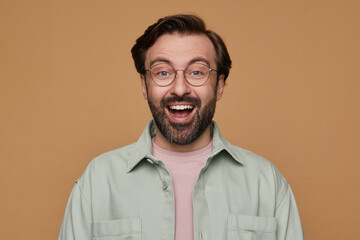 studio portrait of bearded man posing over beige background looking into camera with shocked facial expression and broad smile on his face