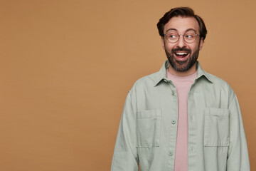 studio portrait of bearded man posing over beige background looks aside with shocked facial expression and broadly smiling