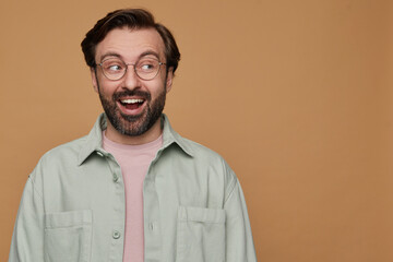 studio portrait of bearded man posing over beige background looks aside with shocked facial expression and broadly smiling