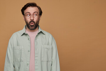 studio portrait of bearded man posing over beige background looks aside with shocked facial expression