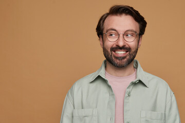 studio portrait of bearded man posing over beige background looks aside with positive facial expression