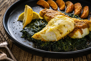 Fish dish - roast cod fillet with potatoes and spinach served on stone plate on wooden table
