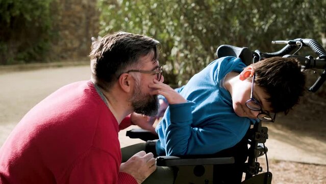 A father having fun with his son with disability in a wheelchair by tickling him while they enjoy time together outdoors.