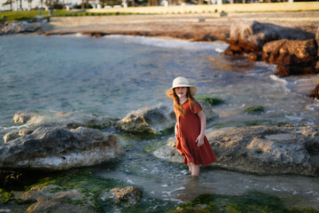 child in a dress and a hat walks barefoot along the seashore, a girl walks along the water's edge, rest and tranquility on the sea coast, rocks and red stones