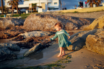 Girl baby toddler in a dress plays on the seashore sunset, lifestyle